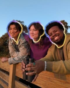 Three friends enjoying sunset view while holding binoculars outdoors.